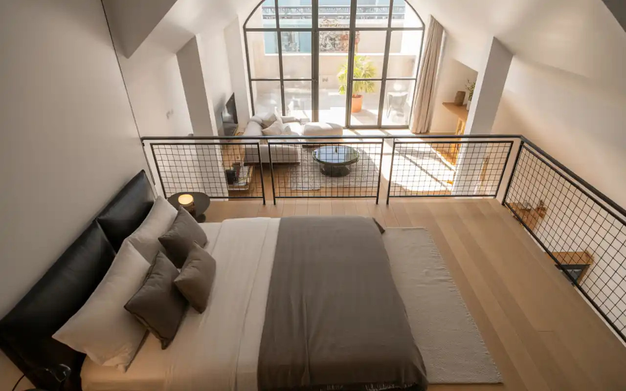 A modern mezzanine loft bedroom with a black metal railing overlooking a bright living room.