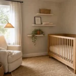 A small gender neutral nursery with warm greige walls, raw oak corner crib, cream linen glider, and floating shelves