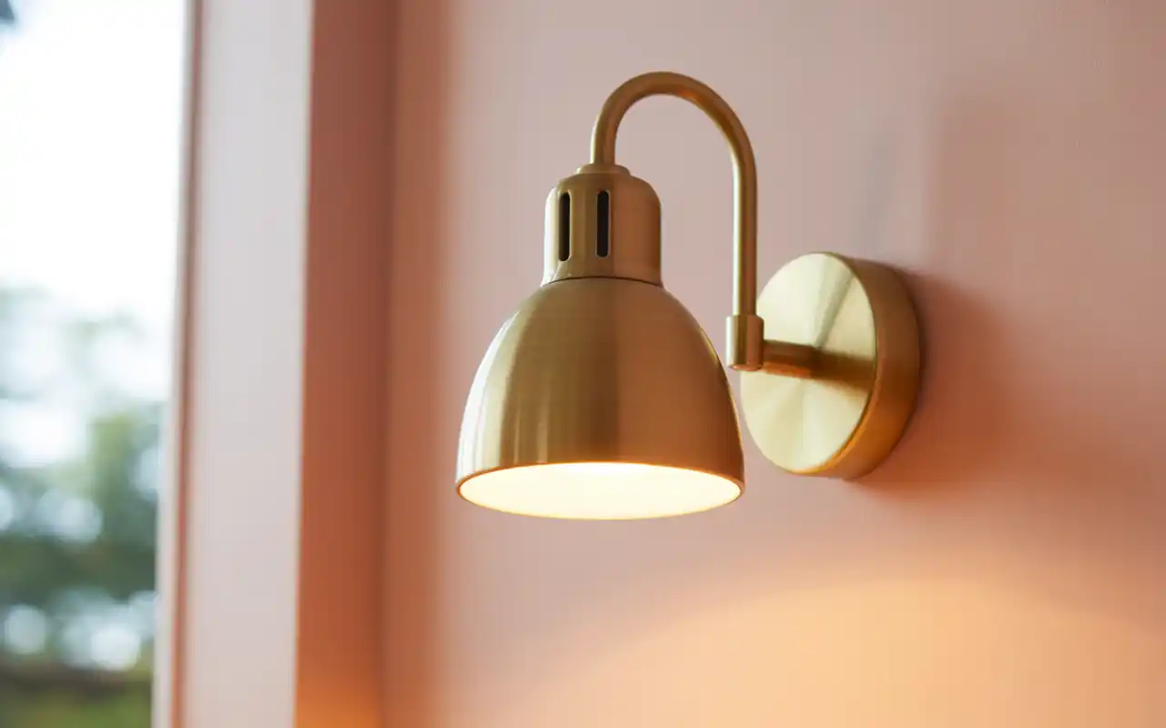 Brushed brass plug-in wall sconce mounted against a warm pale pink bedroom wall.