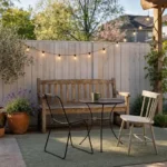 Styled spring backyard with a teak bench, terracotta planters, warm string lights, and a sage green outdoor rug