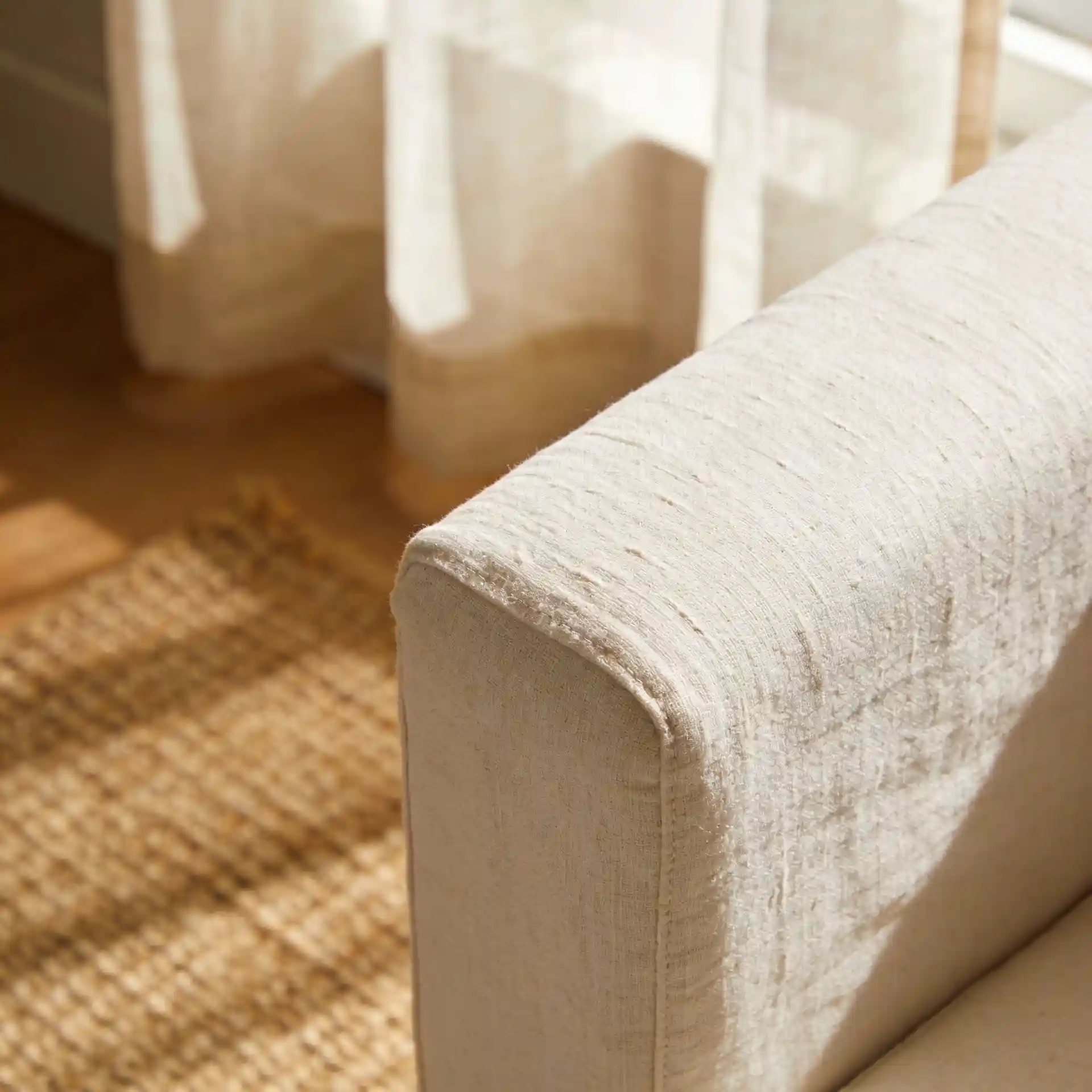 Close-up of cream linen slipcover sofa arm texture with natural jute rug weave and sheer linen curtains in background