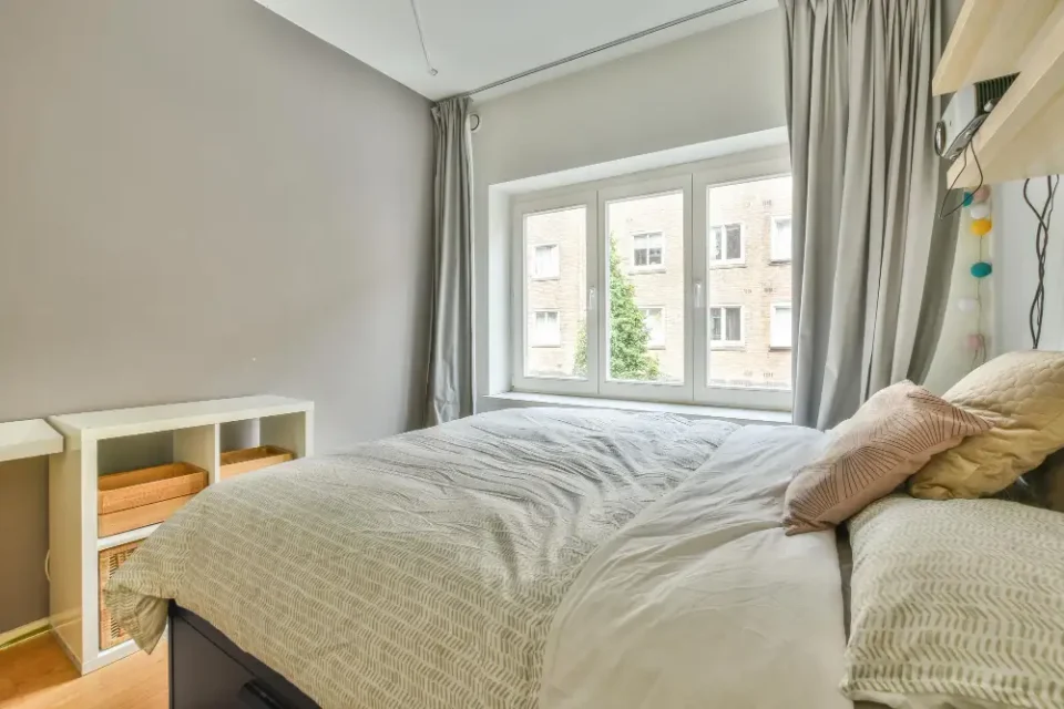 Bright bedroom with a single window, bed on the side wall, sheer curtains, and a large mirror reflecting daylight