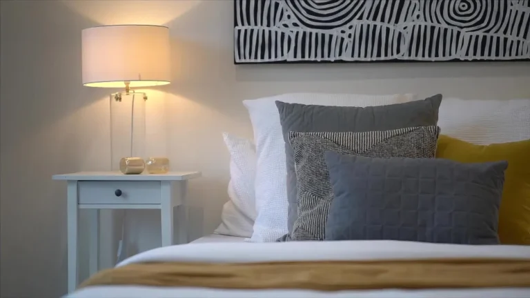 Bright airy white bedroom with warm white walls, layered linen bedding, oak nightstand, and soft 2700K lamp glow