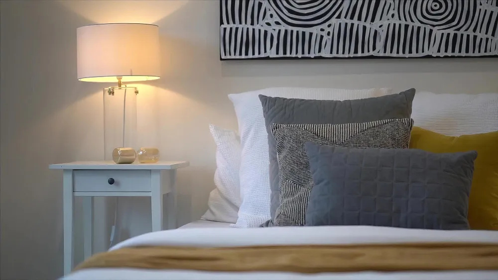 Bright airy white bedroom with warm white walls, layered linen bedding, oak nightstand, and soft 2700K lamp glow