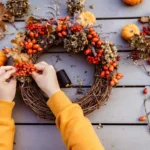 Overhead view of hands wiring rosehips, dried hydrangea, oak leaves and mini pumpkins onto a grapevine wreath on grey boards