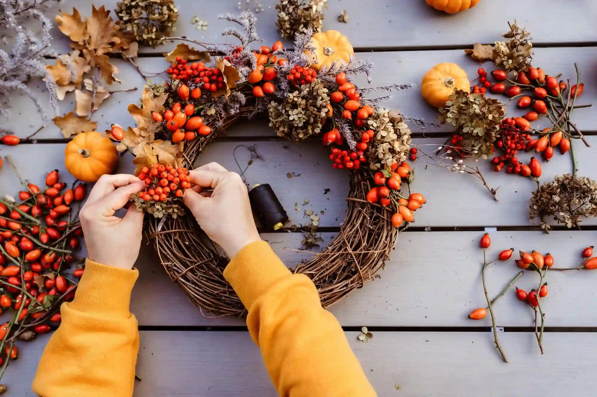 Overhead view of hands wiring rosehips, dried hydrangea, oak leaves and mini pumpkins onto a grapevine wreath on grey boards